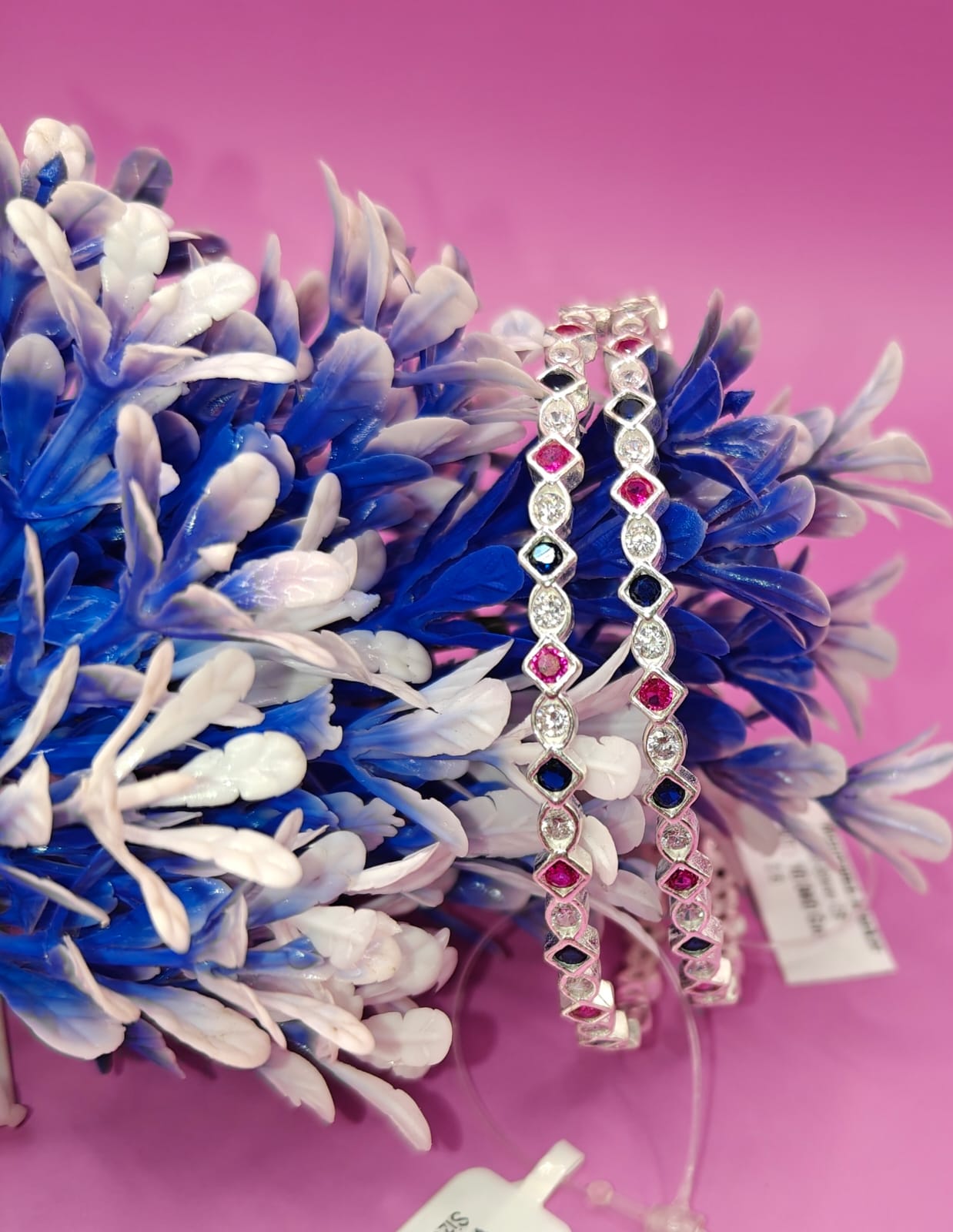 Two silver-toned beaded bracelets with alternating red and dark blue gemstones, displayed on a pink background next to blue and white decorative flowers.