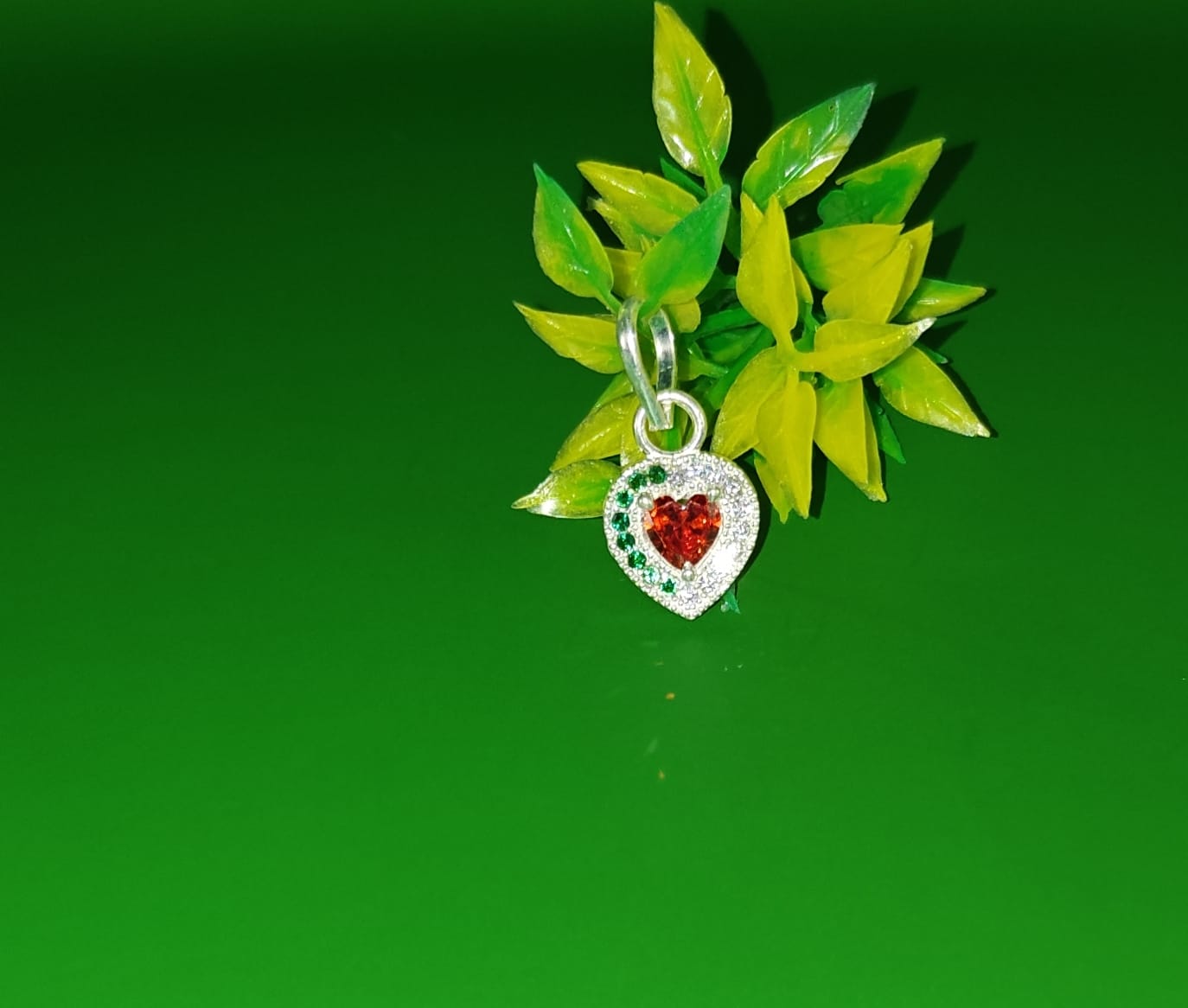 A silver heart-shaped pendant charm with a red heart in the center and small green and clear stones around the border