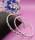 Two silver-toned bangles with black and pink floral engravings, displayed on a black velvet stand against a pink background with blurred blue and white flowers.