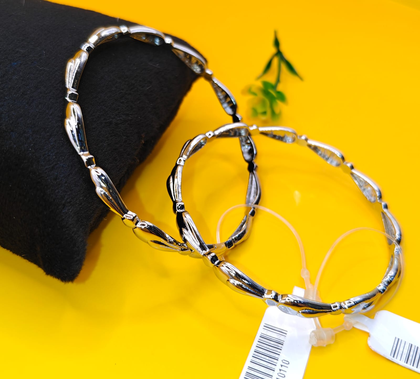 Two silver-toned twisted bangles displayed on a bright yellow surface next to a black fabric cushion, with a small green sprig and a white product tag visible.