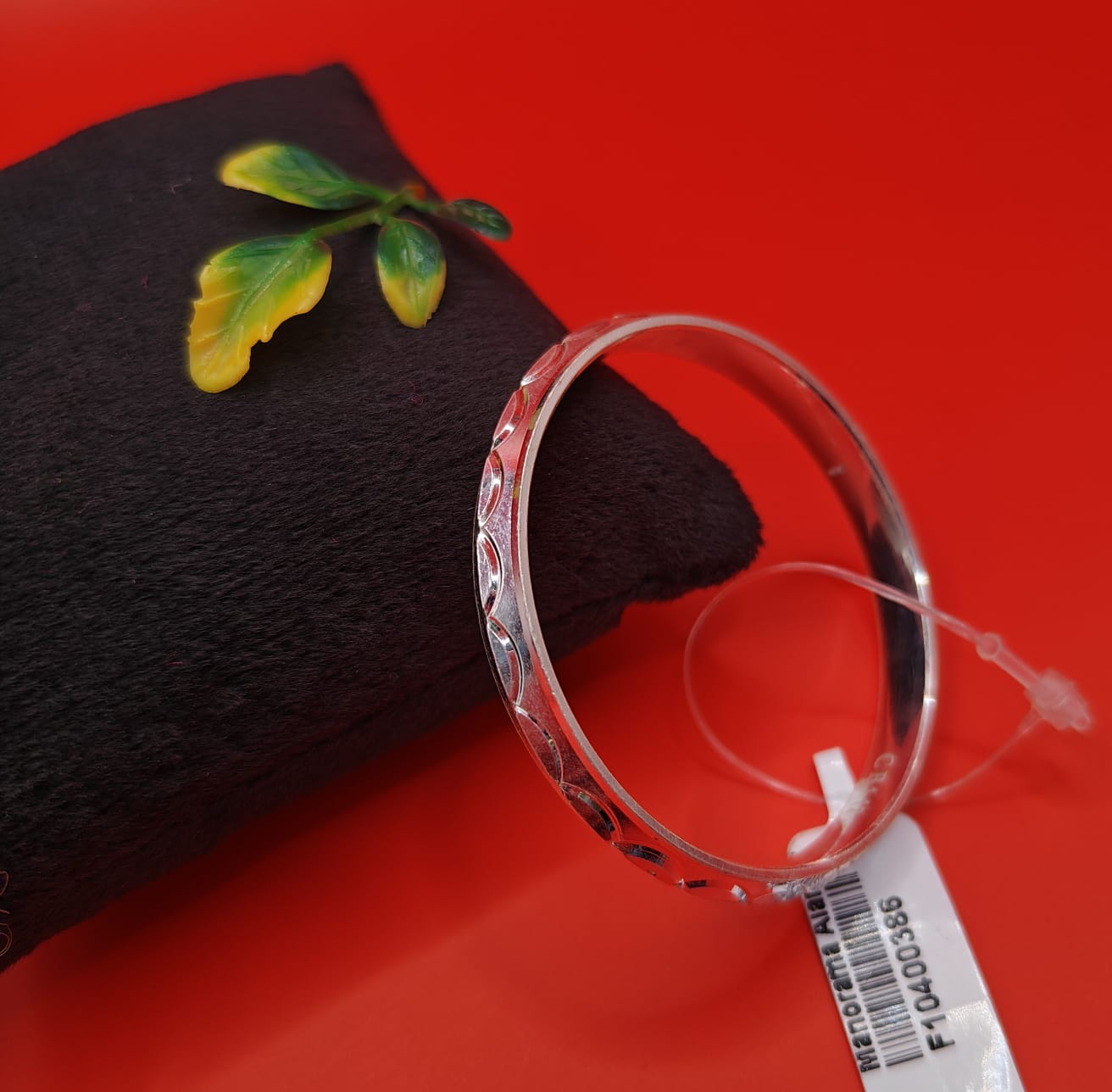 A clear glass bangle bracelet with a red patterned border, resting on a black textured cushion with a small green leaf decoration, set against a solid red background.