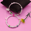 Two silver beaded bracelets with small multicolor accent beads, displayed on a pink background with a black cushion and a yellow flower prop.