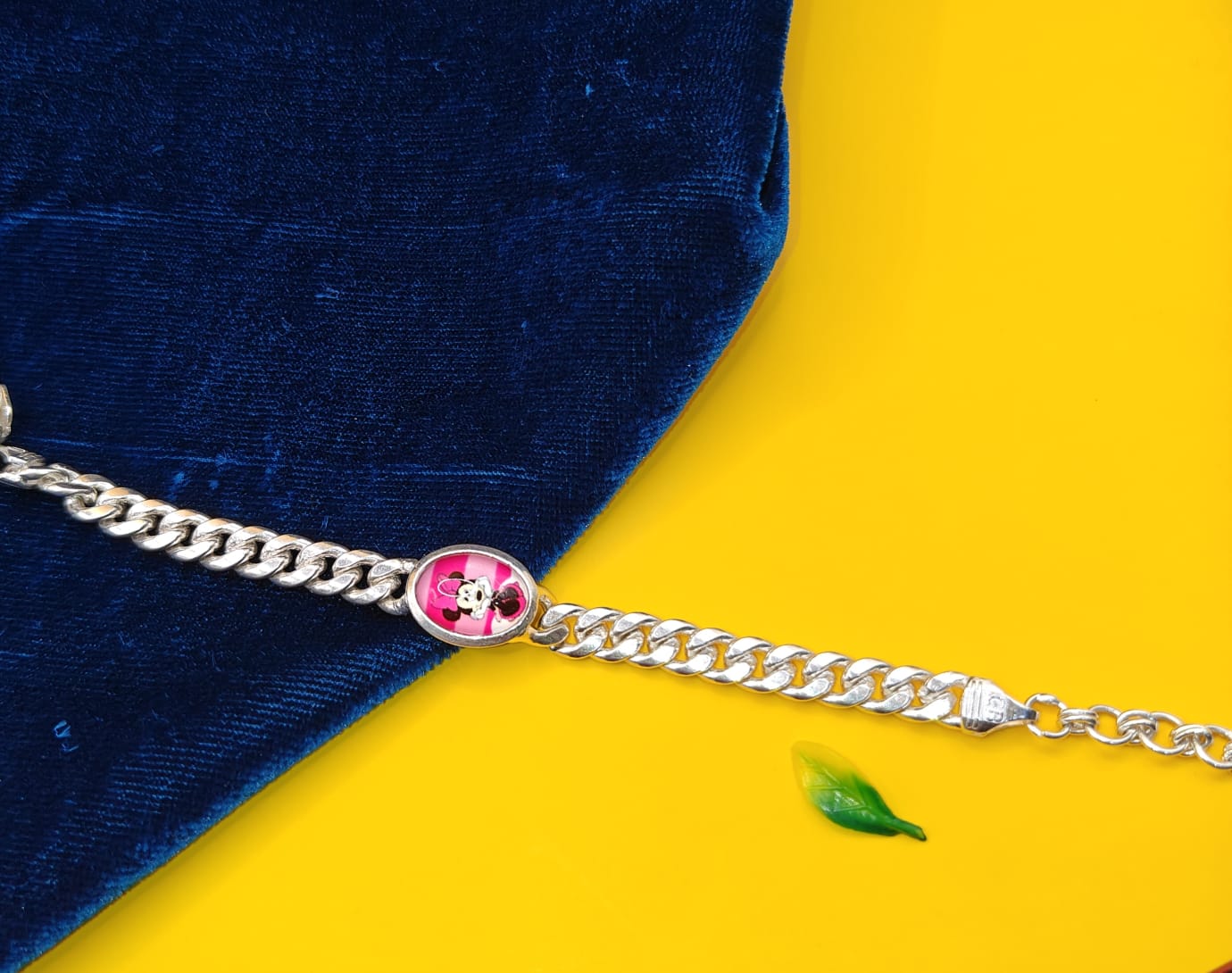 A silver-toned curb chain bracelet featuring a round pink and white Minnie Mouse charm, displayed diagonally across a split background of dark blue velvet and bright yellow.