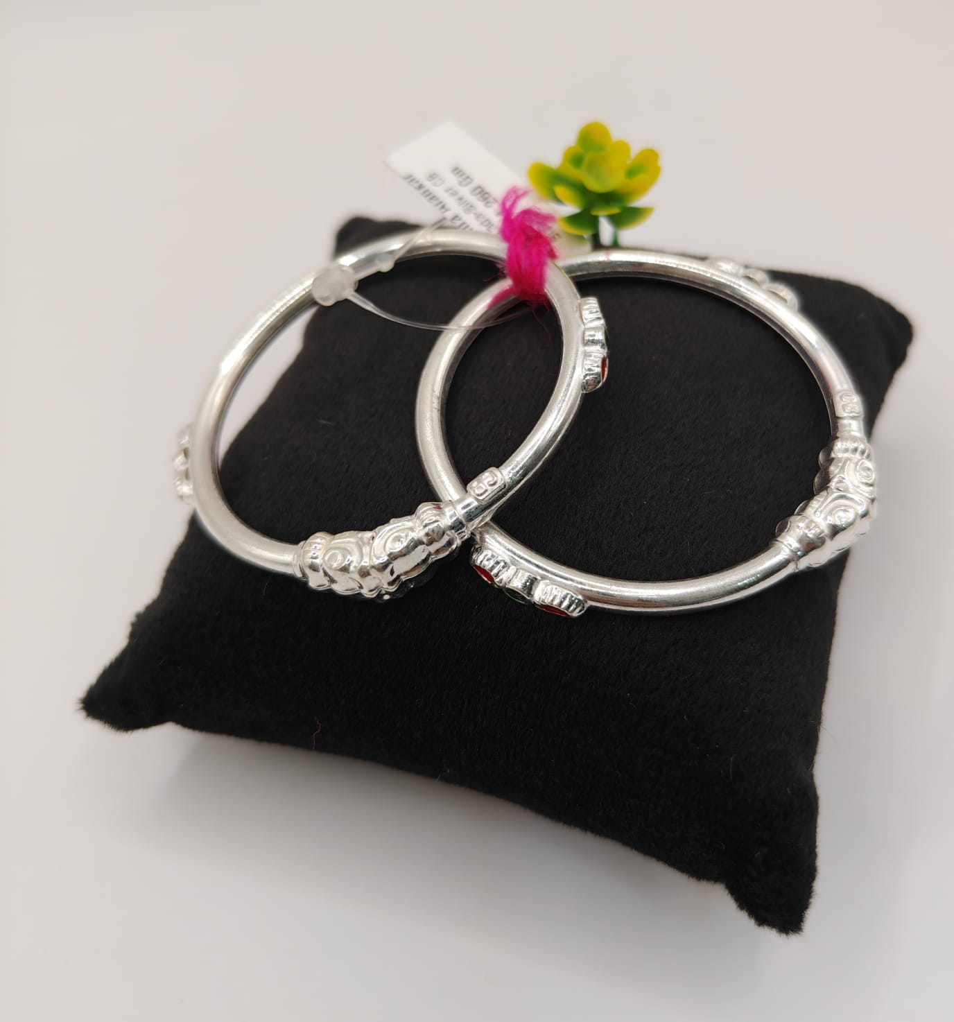 A pair of silver-toned bangles with decorative textured ends, displayed on a black velvet cushion with a small pink flower and tag.